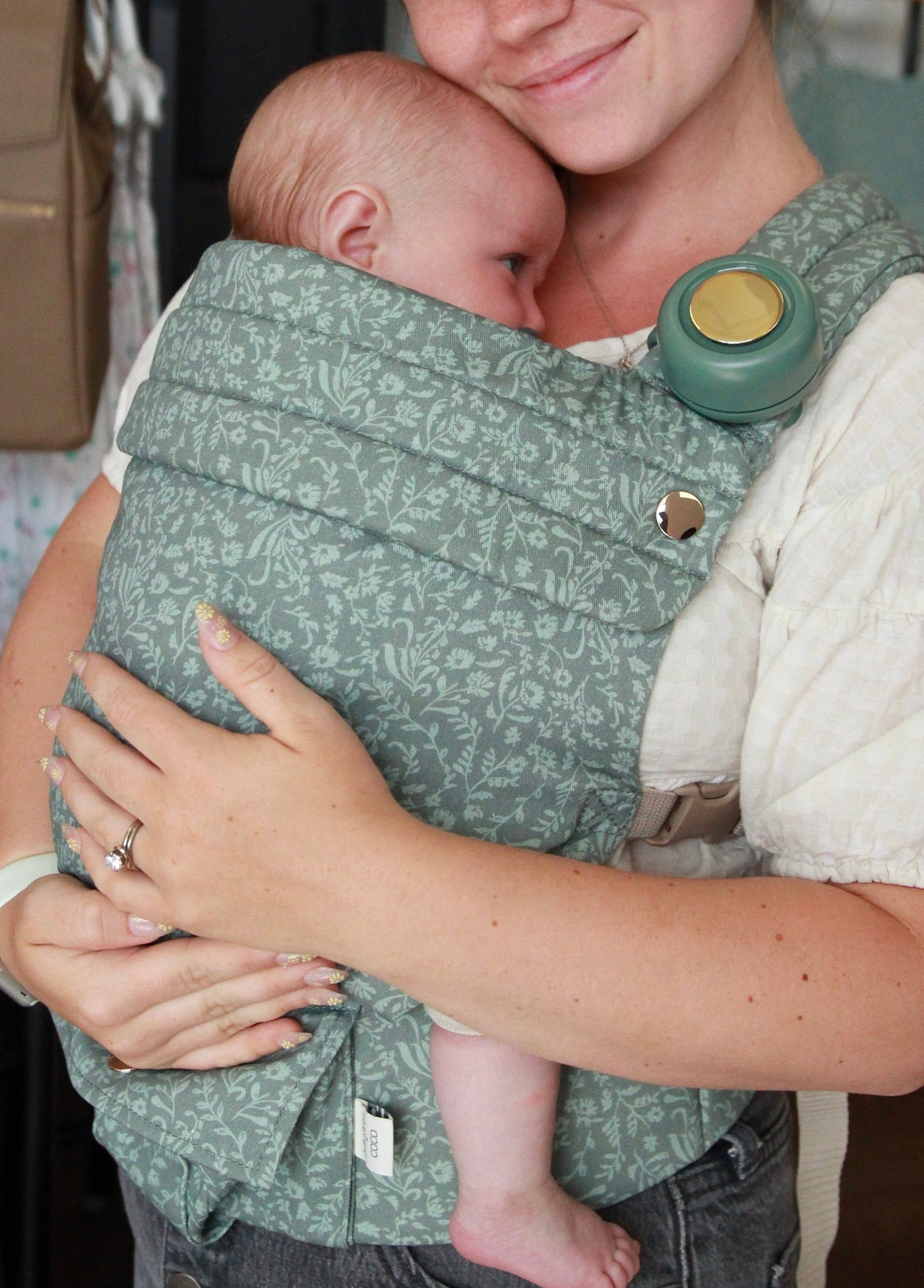 Teal floral baby carrier with pocket and round button detail, newborn resting inside