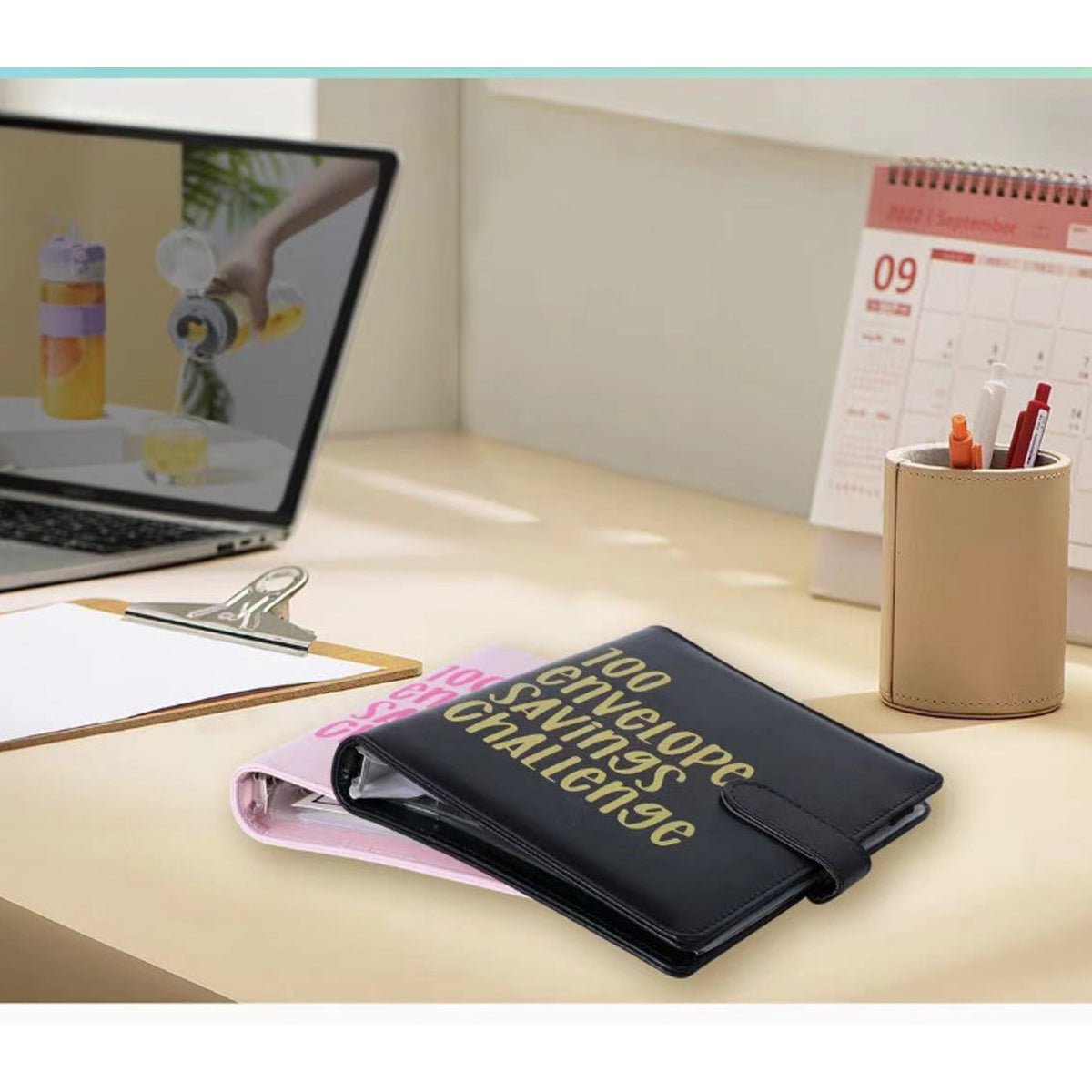 Black and pink envelope savings challenge binders on a desk with pens and calendar.
