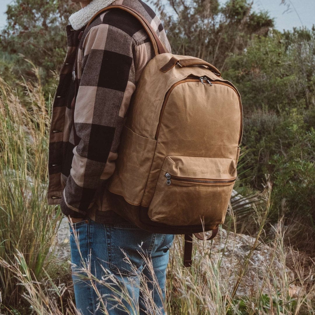 Tan canvas backpack with front zip pocket and brown shoulder straps, worn