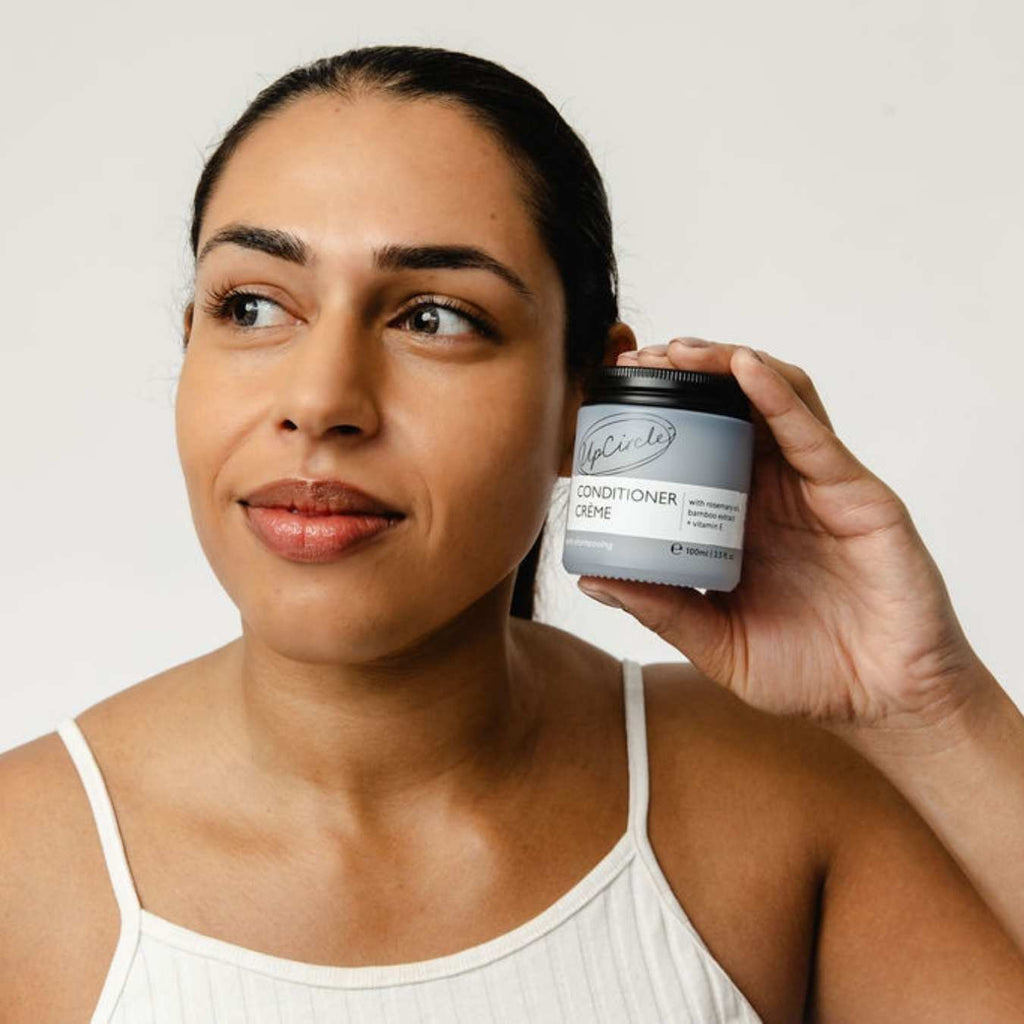 UpCircle shampoo creme jar held beside face, light blue jar with black lid.