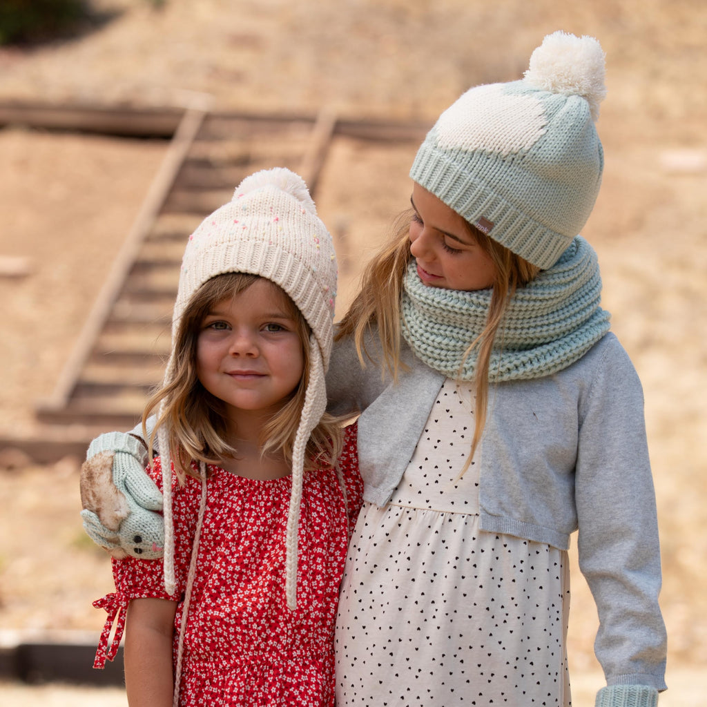 Two kids wearing off-white knit earflap beanies with pom-poms and tie strings.