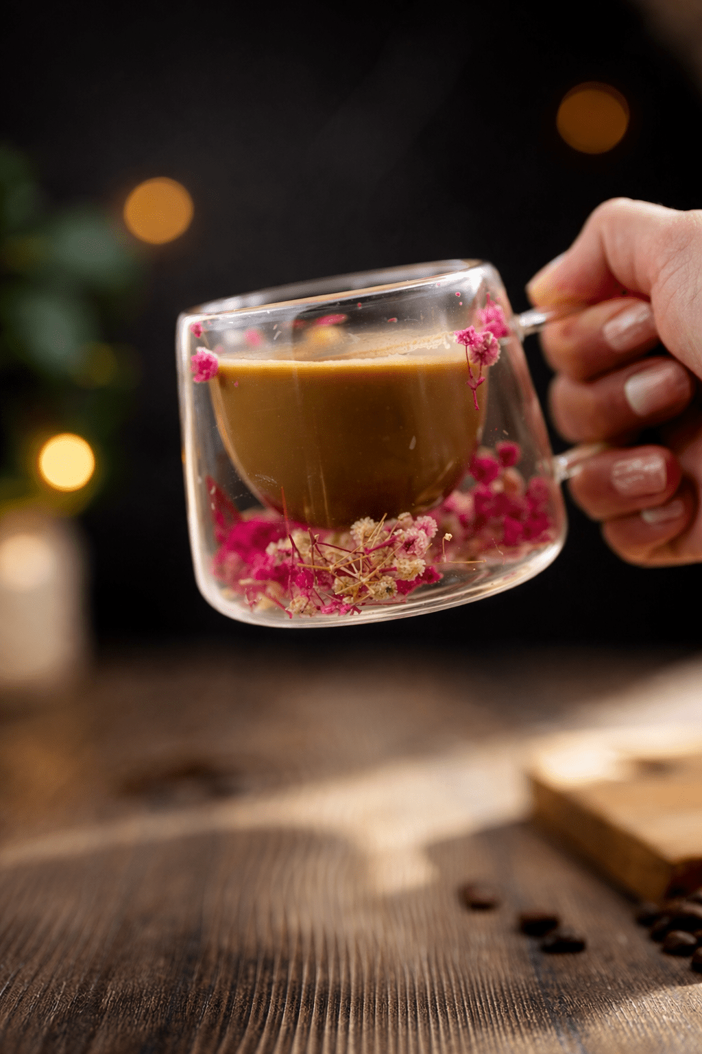 Hand holding floral glass mug with pink dried flowers in dark tea, clear handle