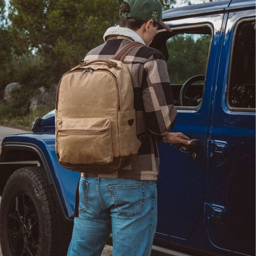 Sand waxed canvas backpack worn by a person near a blue vehicle, side view