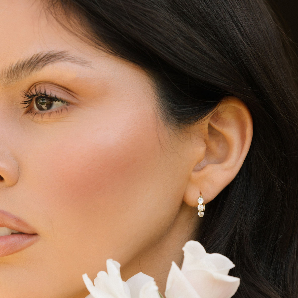 Gold-tone huggie hoop earrings with three clear stones, ear profile close-up