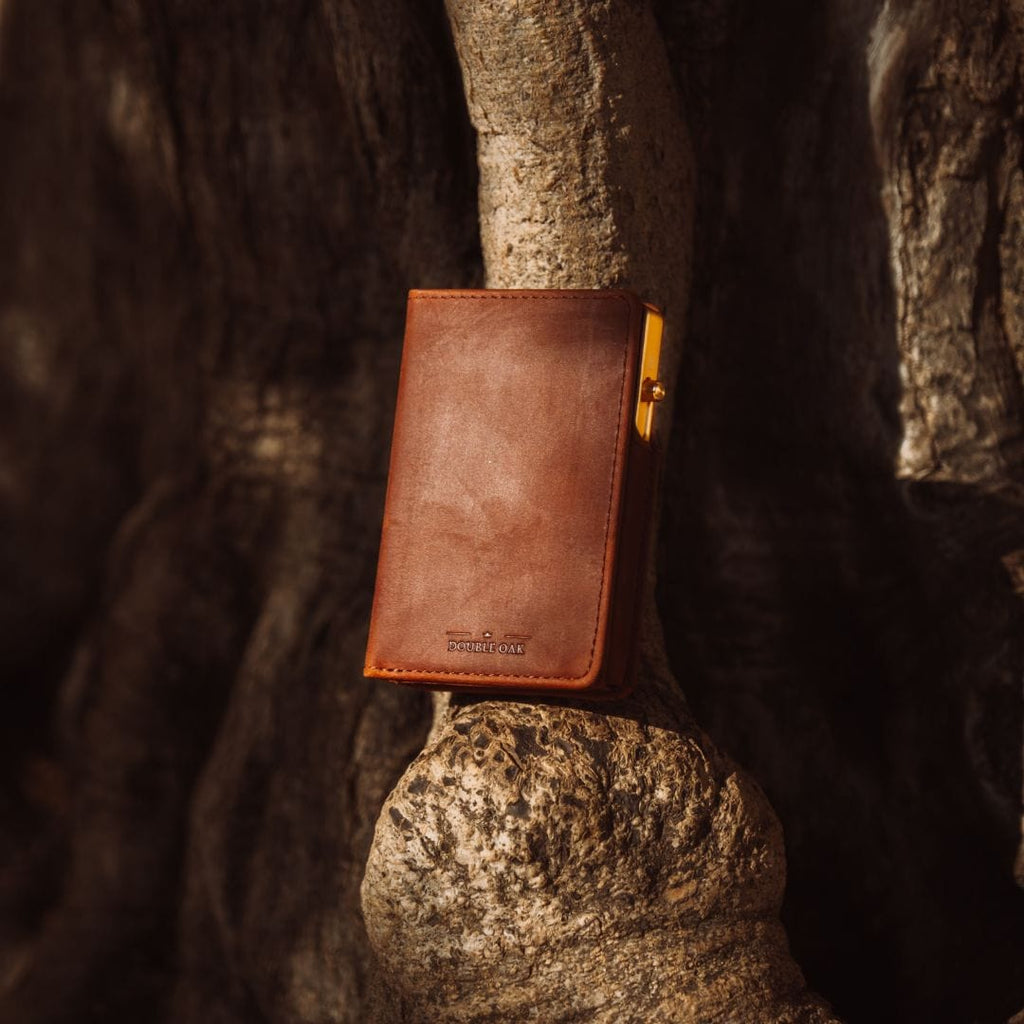 Tan leather bifold wallet with gold clasp and stitched edges, standing on stone.