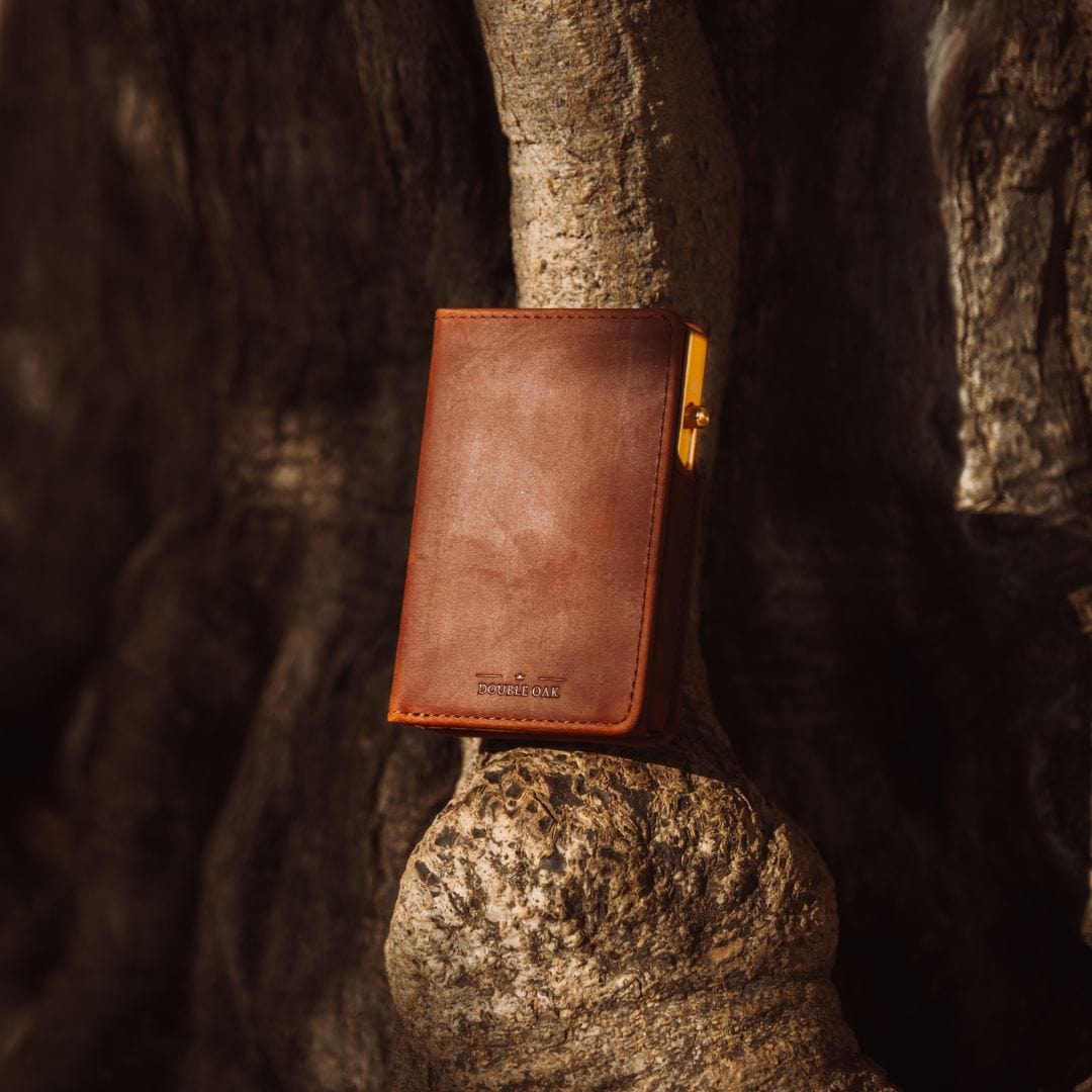 Tan leather bifold wallet with gold clasp and stitched edges, standing on stone.