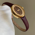 Ladies gold-tone watch with crystal bezel, dark dial, burgundy leather strap.
