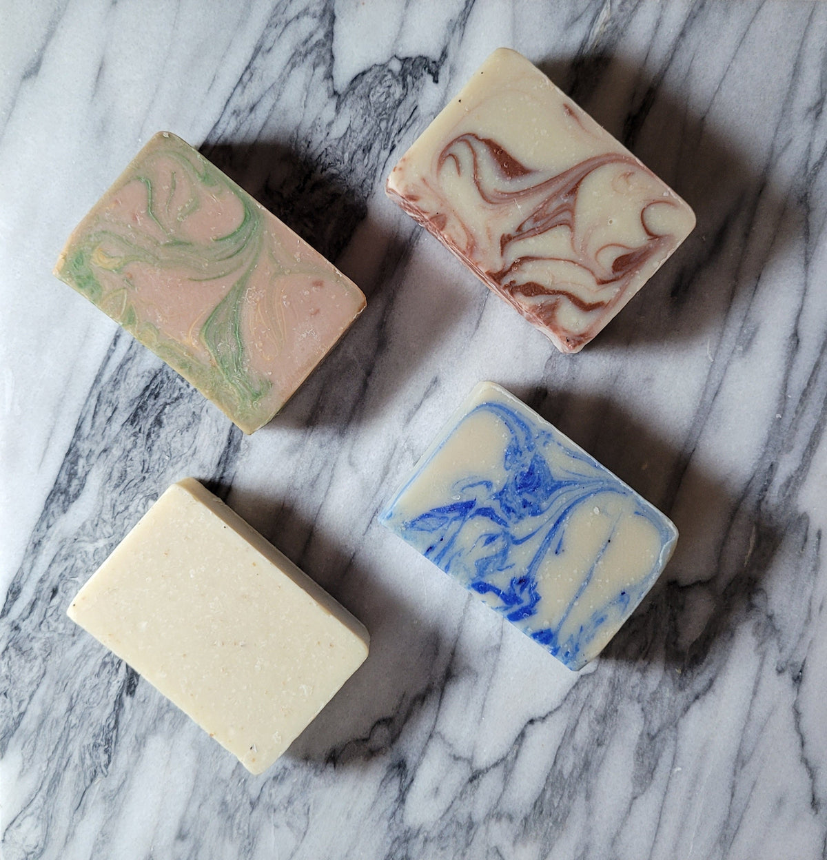 Assorted cold-processed soap bars on marble: green, red-swirl, blue, and beige.
