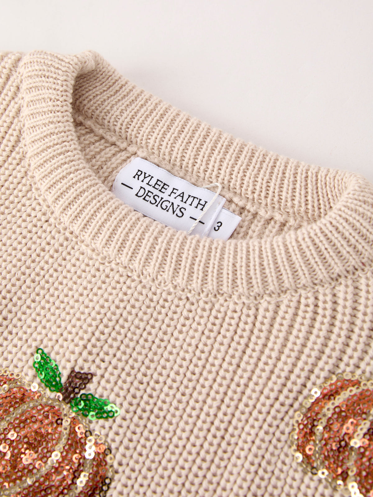 Beige knit sweater neckline with RYLEIGH FAITH DESIGNS tag and pumpkin sequins
