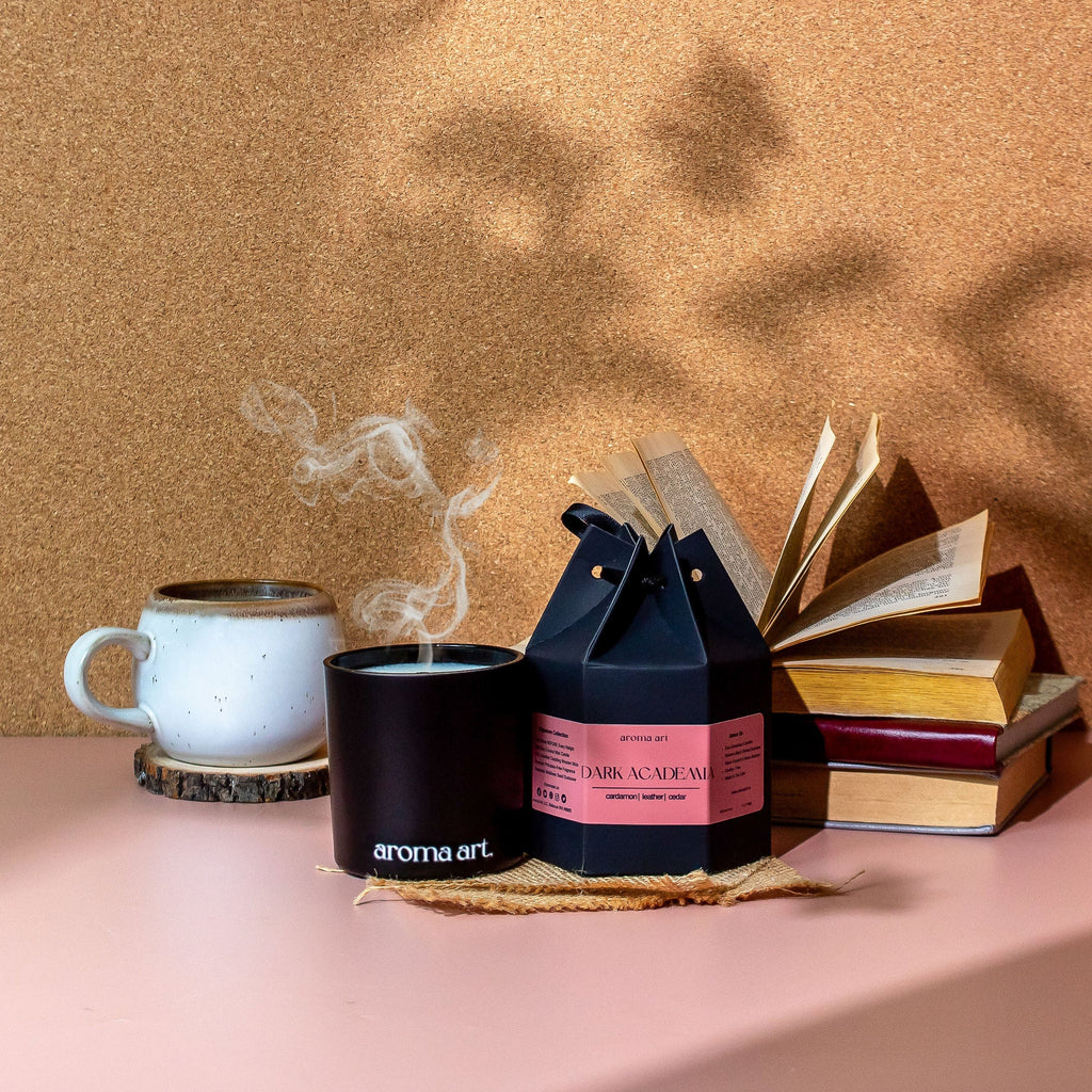 Black wood wick candle with lit flame, beside boxed candle and small books
