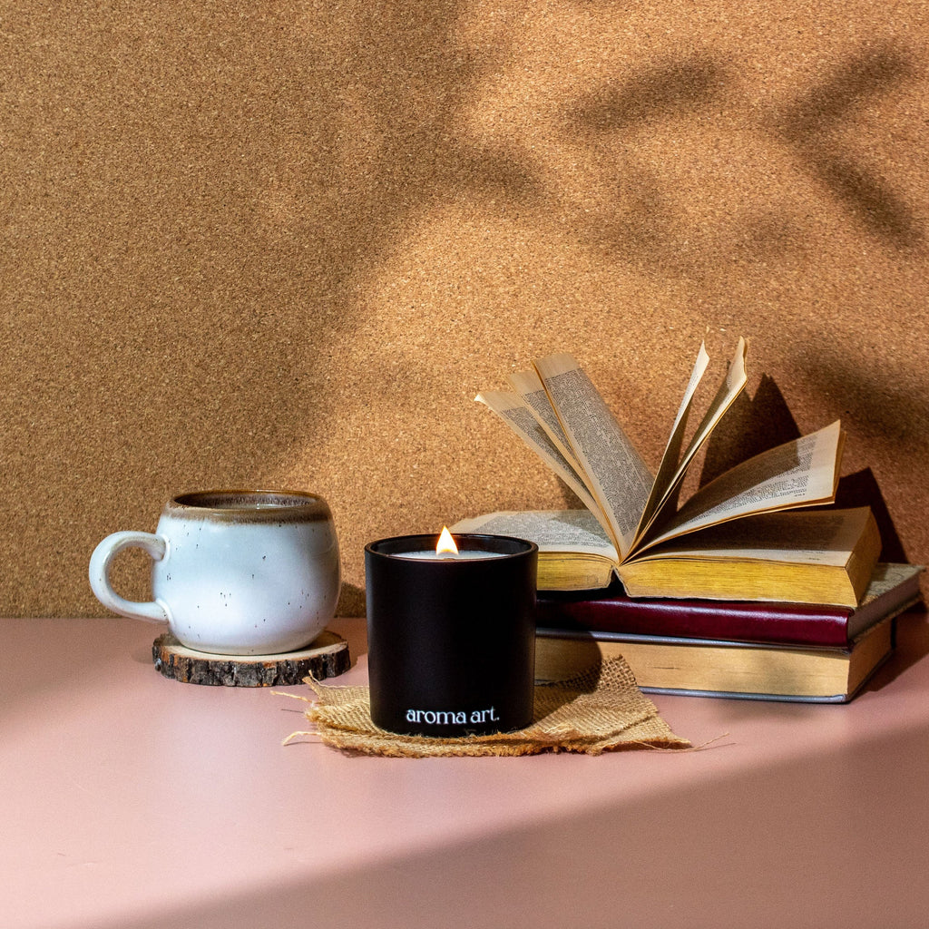 Black wood wick candle with flame next to scented books and a ceramic mug