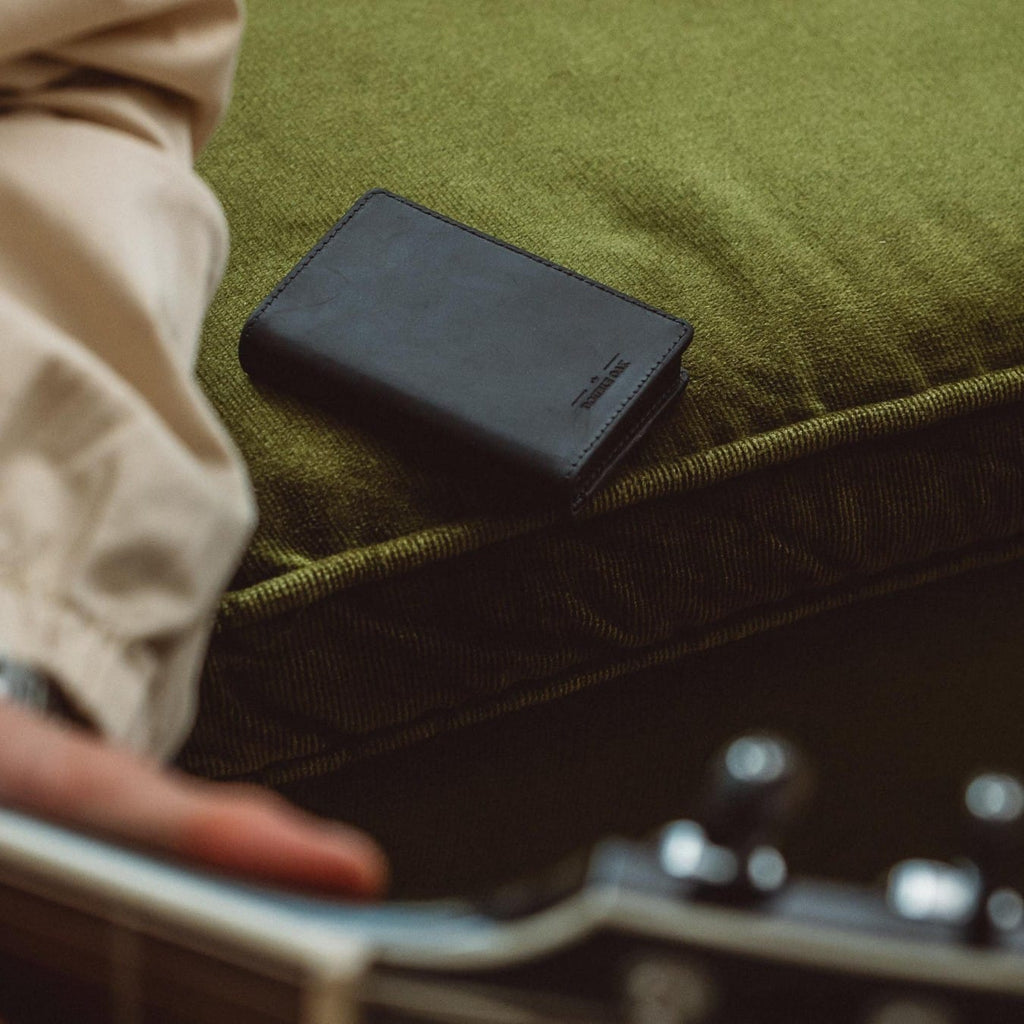 Bifold wallet, dark leather, resting on green fabric with logo on the front panel.