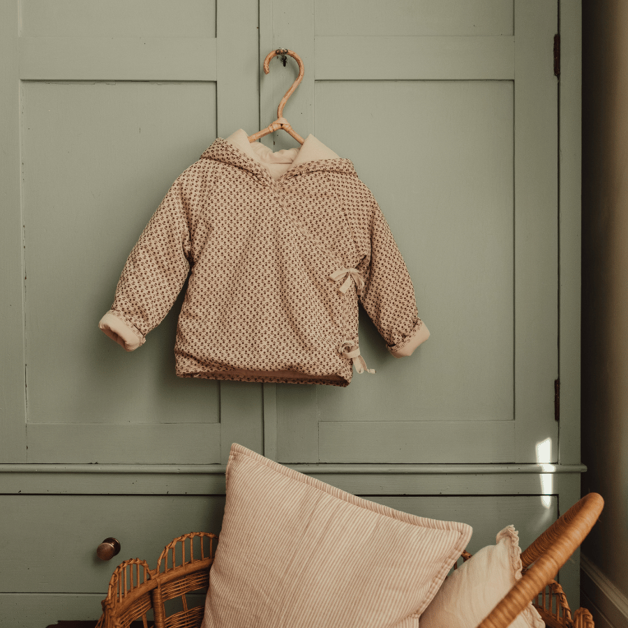 Light pink hooded baby jacket with allover brown print, hanging on a wooden hook