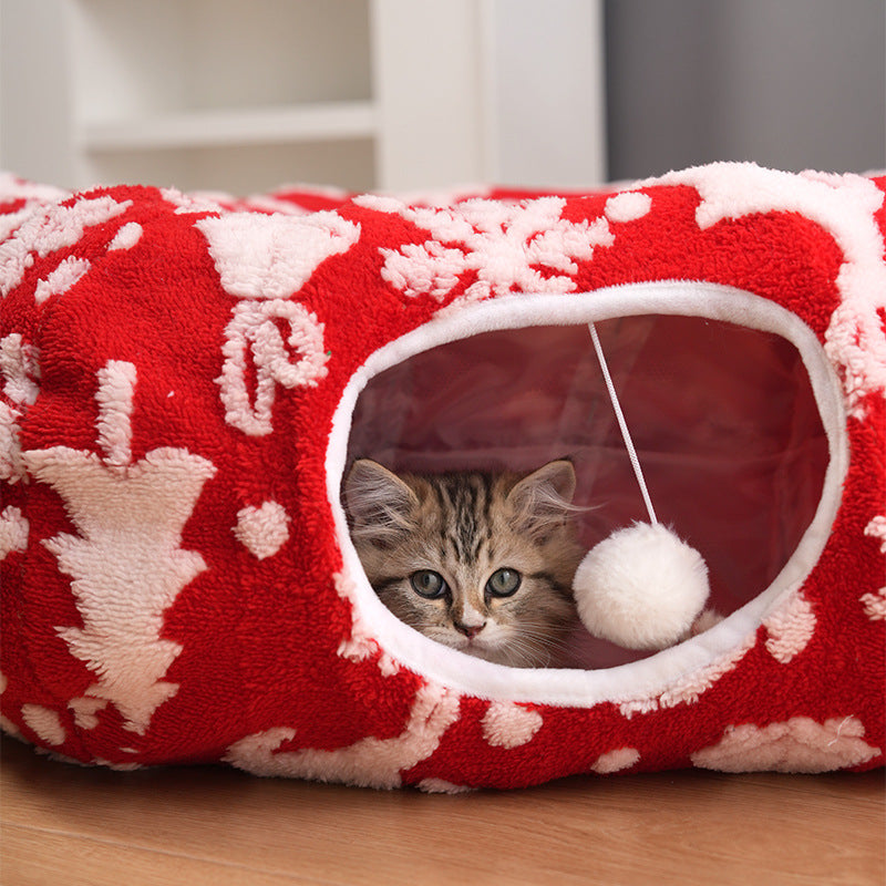 Red plush pet bed with white snowflake print and oval tunnel opening, kitten peeking out