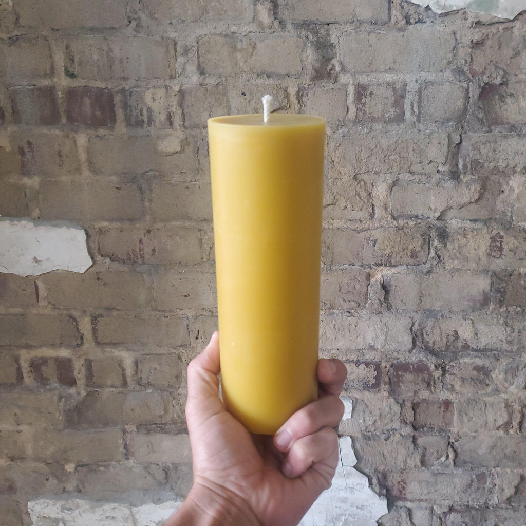 Hand holding a single tall yellow beeswax pillar candle with wick on top