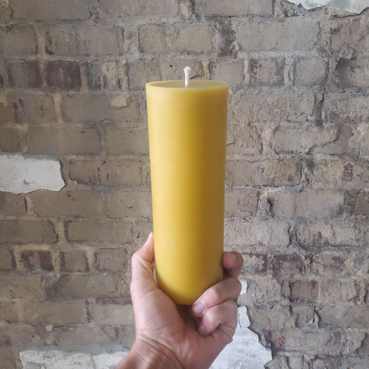 Hand holding a single tall yellow beeswax pillar candle with wick on top