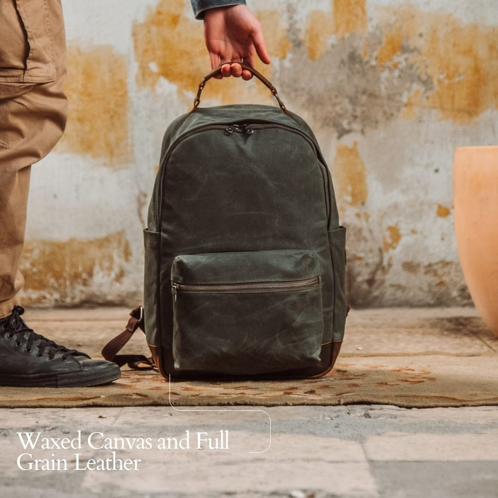 Green waxed canvas backpack held by top handle; front zip pocket and side tabs.