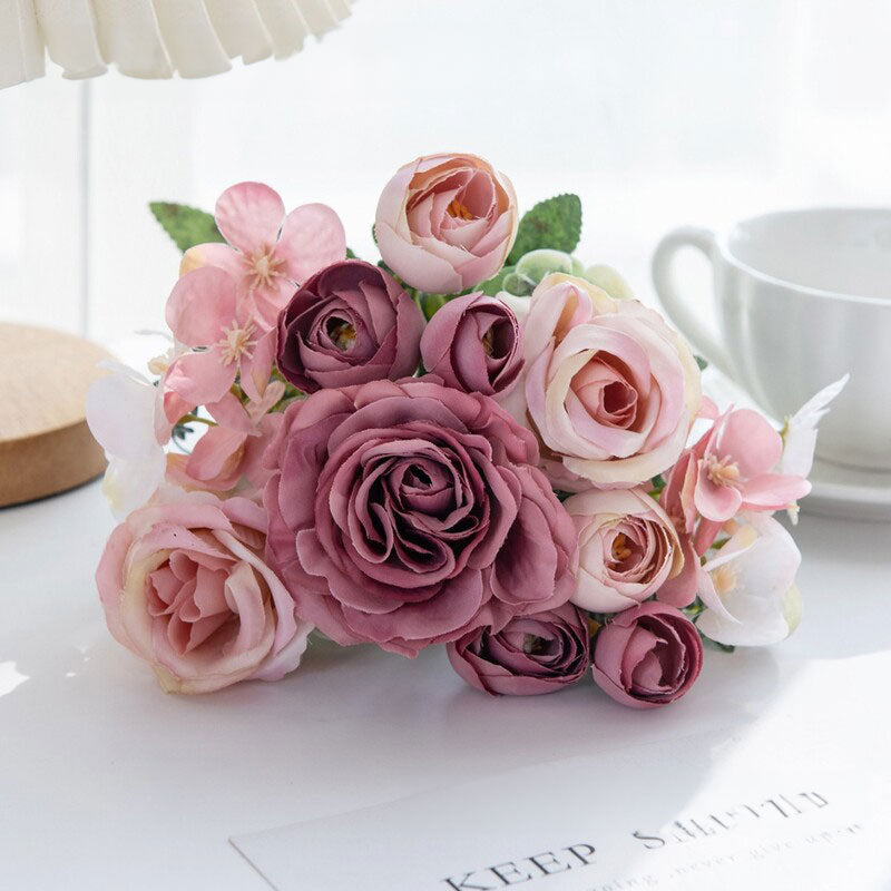Pink and purple silk rose bouquet with small blossoms and green leaves in a clear vase