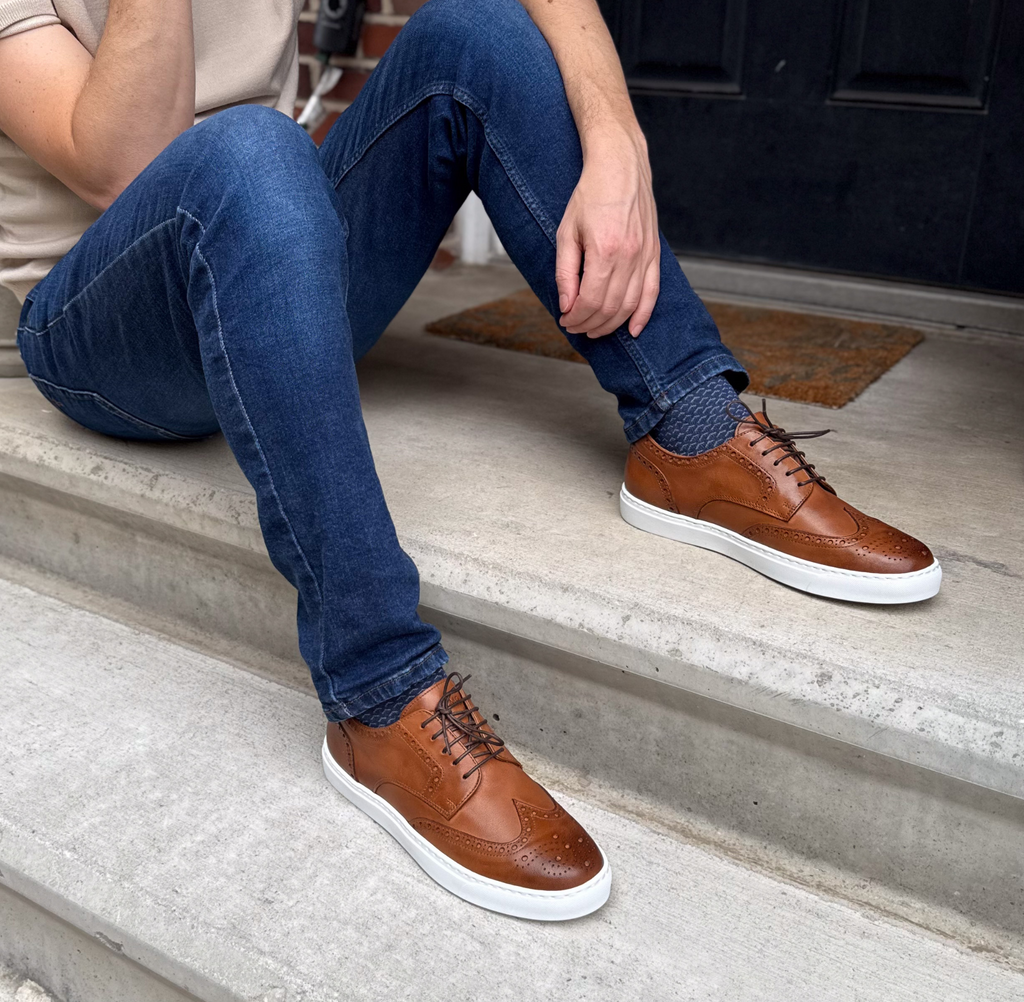 Men's cognac wingtip lace-up shoes with white soles, sitting pose