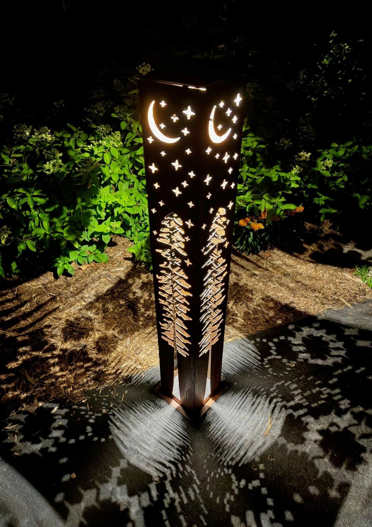 Rust-colored solar light pillar with glowing moon, stars, and pine tree cutouts at night.