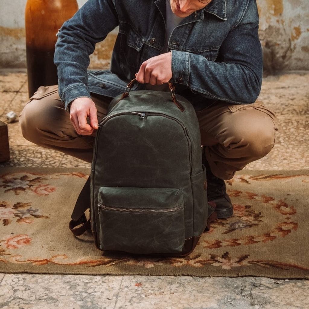 Green canvas backpack on floor with top handle, shoulder straps, and front zip pocket.