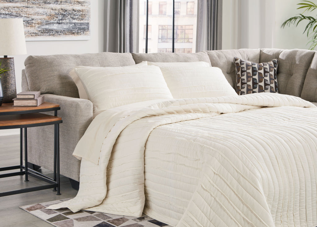 Light gray chaise sleeper sectional with open bed, quilt, and decorative pillows