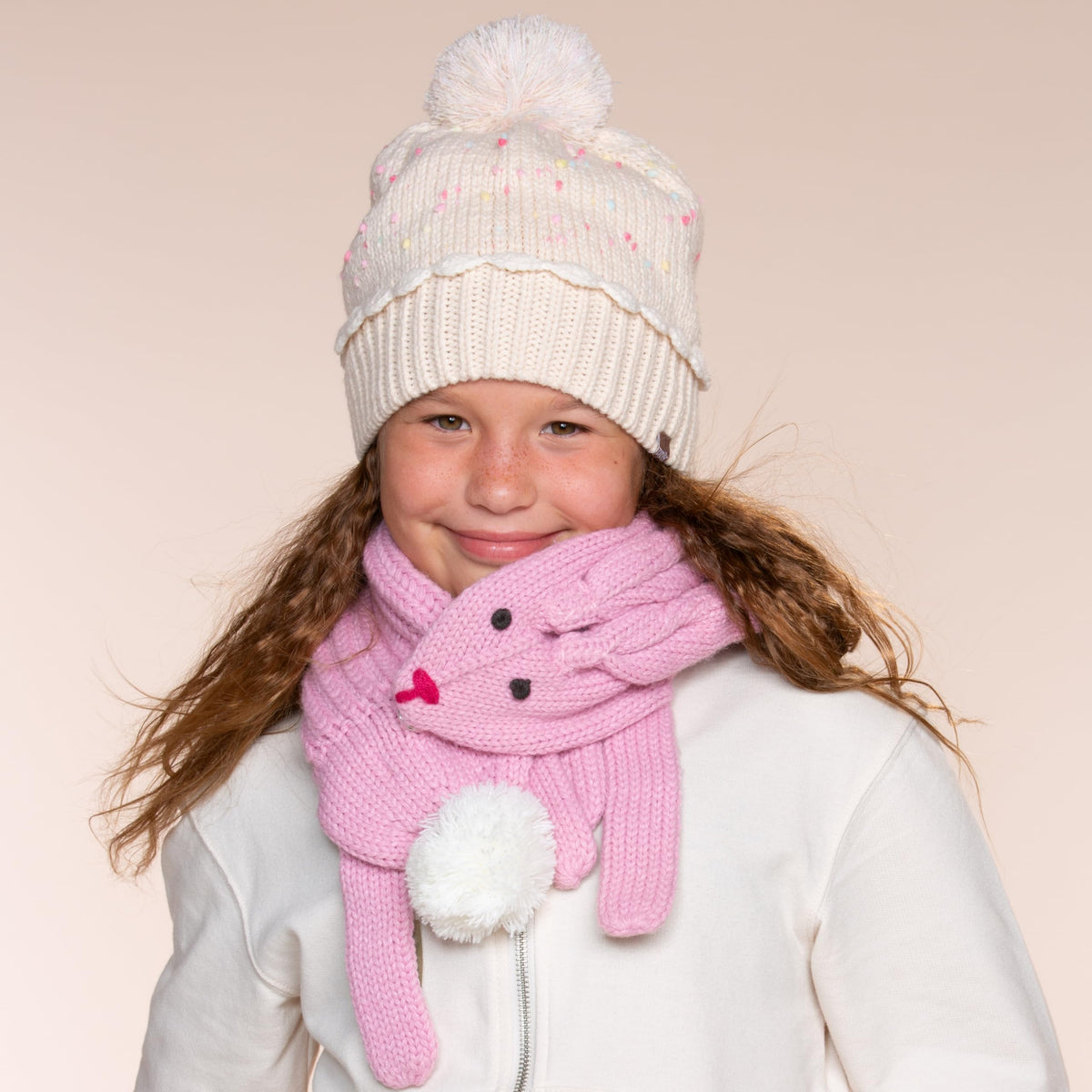 Toddler girl wearing cream speckled beanie hat and pink character mittens with pom