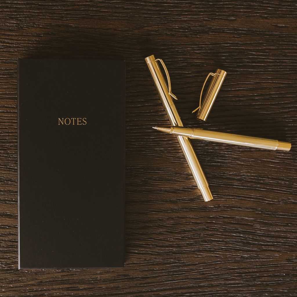 Black notes booklet with gold “NOTES” label next to a gold pen on a desk.