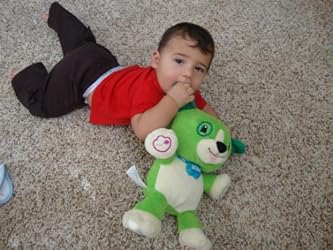 Green plush song-learning bear toy lying on carpet with a baby reaching toward it