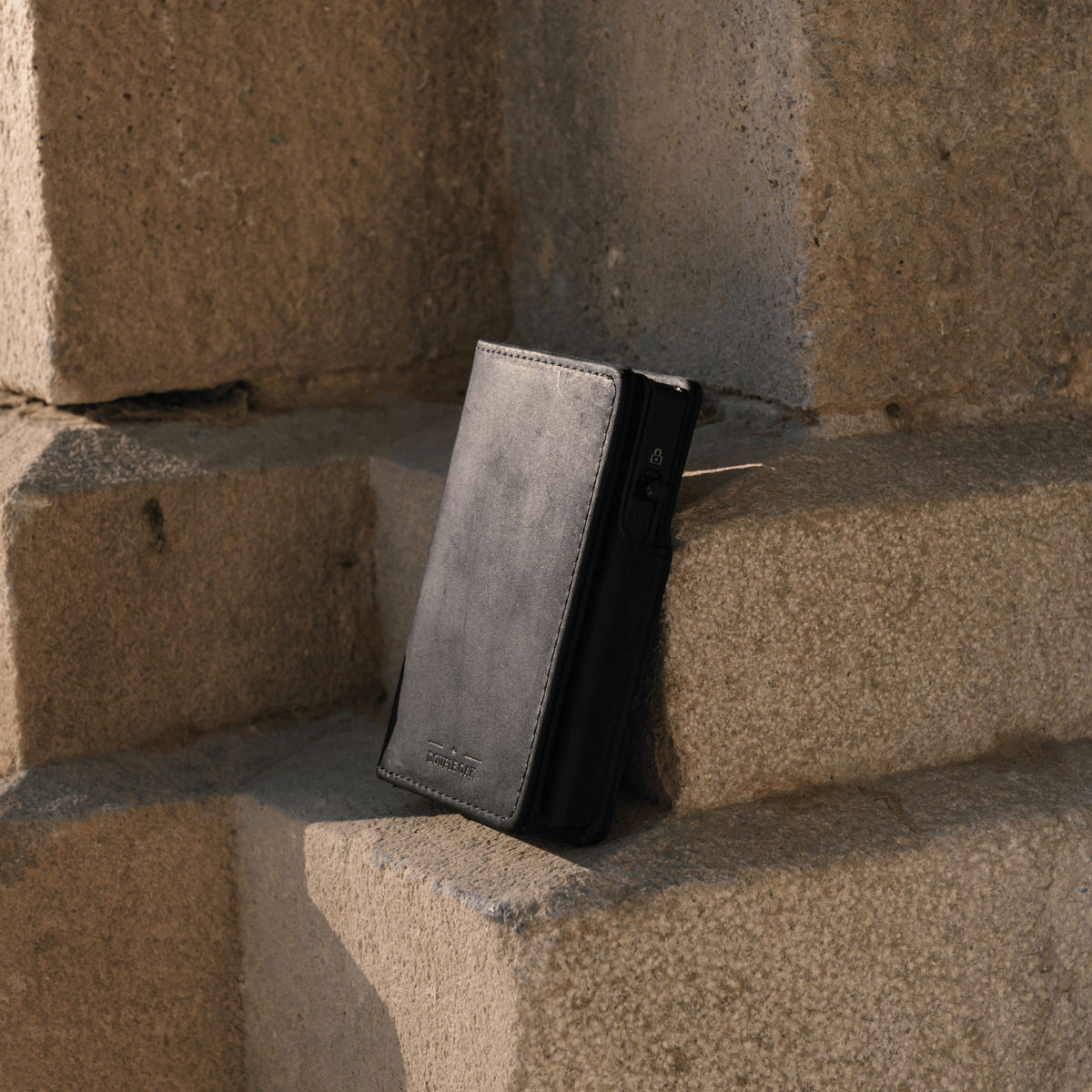 Gray bifold wallet with dark side panel and card slots, standing against bricks.