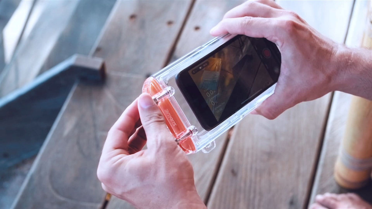 Hands holding a clear waterproof phone case with orange-tinted latch and front window.