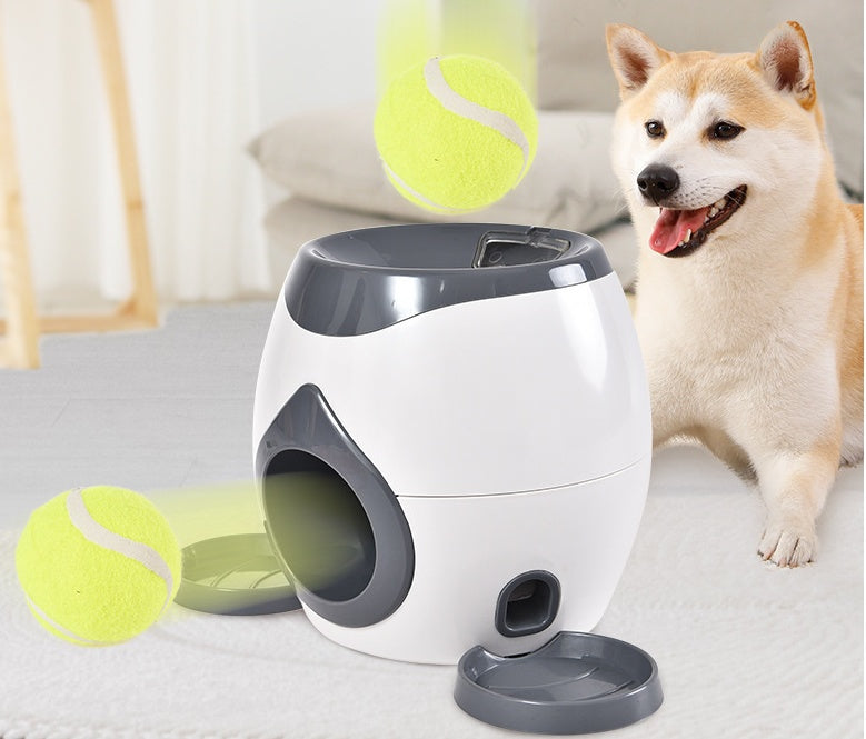 White and gray smart pet feeder with front ball launcher and tennis balls, with dog nearby