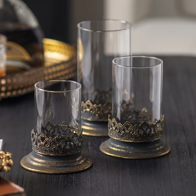 Three glass candle holders with clear cylinders and ornate dark metal band bases
