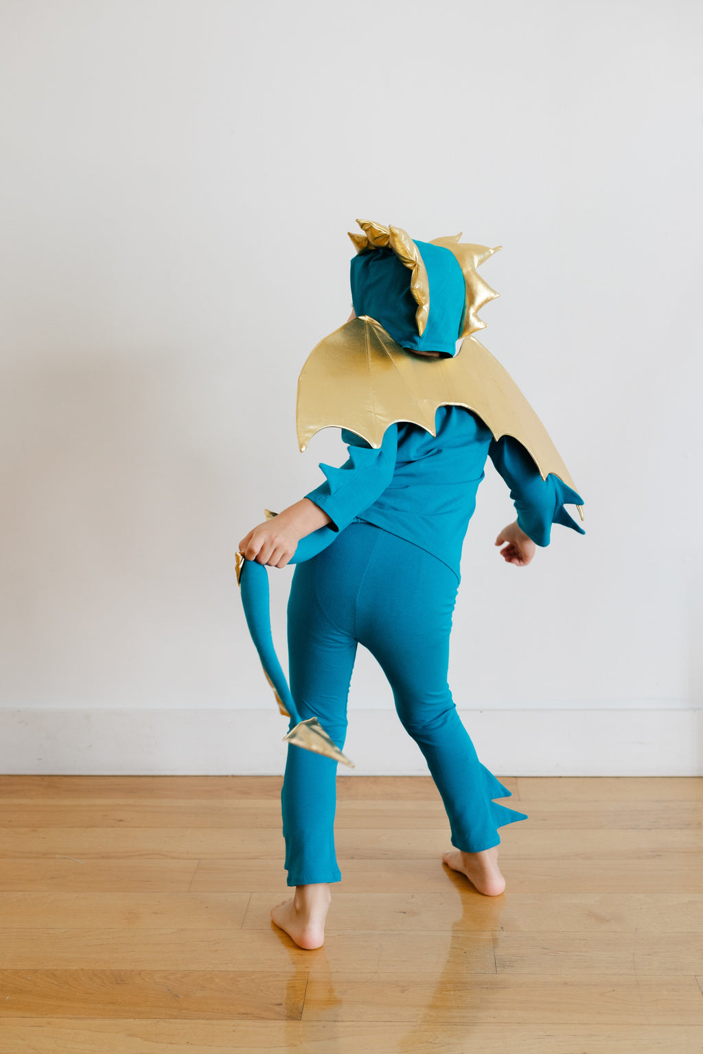 Turquoise dragon costume with gold wings and tail, toddler walking sideways indoors.