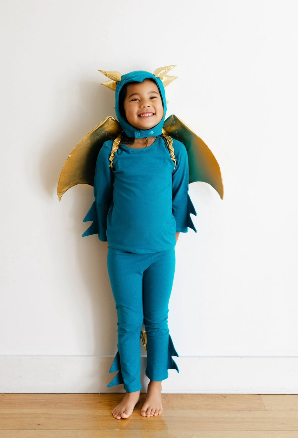 Turquoise dragon costume with gold winged hood, toddler standing barefoot by wall.