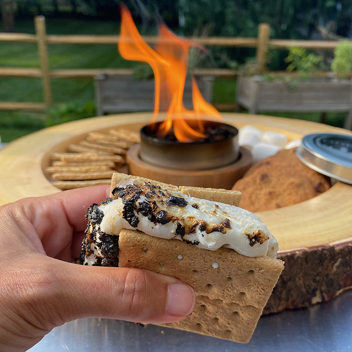 Hand holding a s'more with melted marshmallow and chocolate by a fire pit