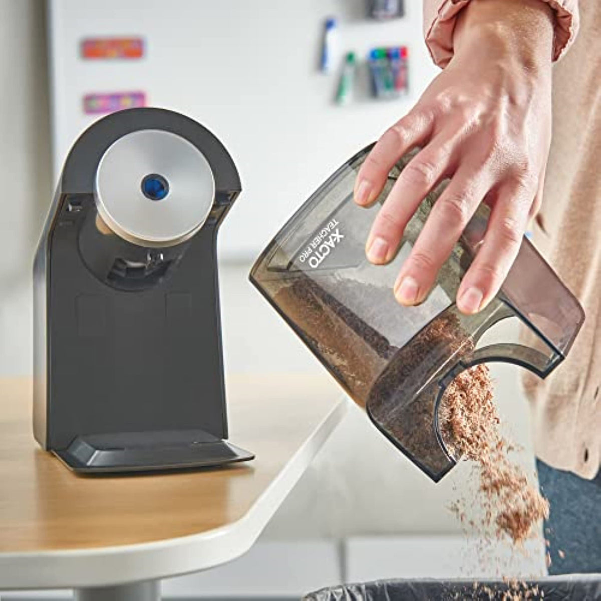 Black electric pencil sharpener with emptying tray being tilted over clear bin with shavings.