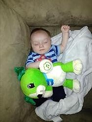 Green plush song-learning bear toy next to a sleeping baby on a bed