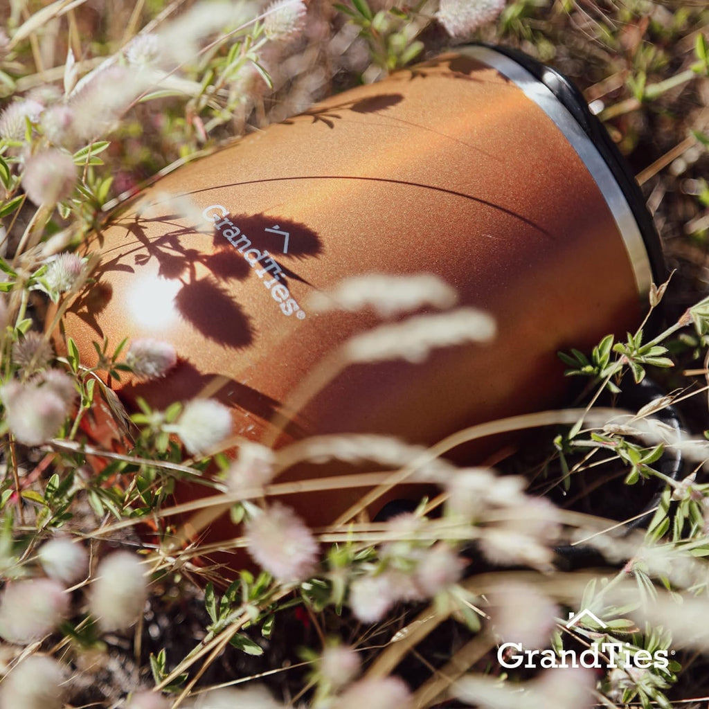 Cognac metallic insulated coffee mug lying in grass, black lid and handle visible