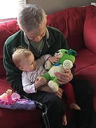 Green plush song-learning bear toy in an adult’s hands with a baby nearby