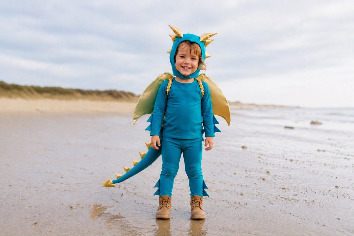 Turquoise dragon costume with gold wings and headpiece, toddler standing on beach sand.
