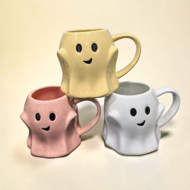 Three ghost-shaped ceramic mugs in yellow, pink, and white with smiling faces