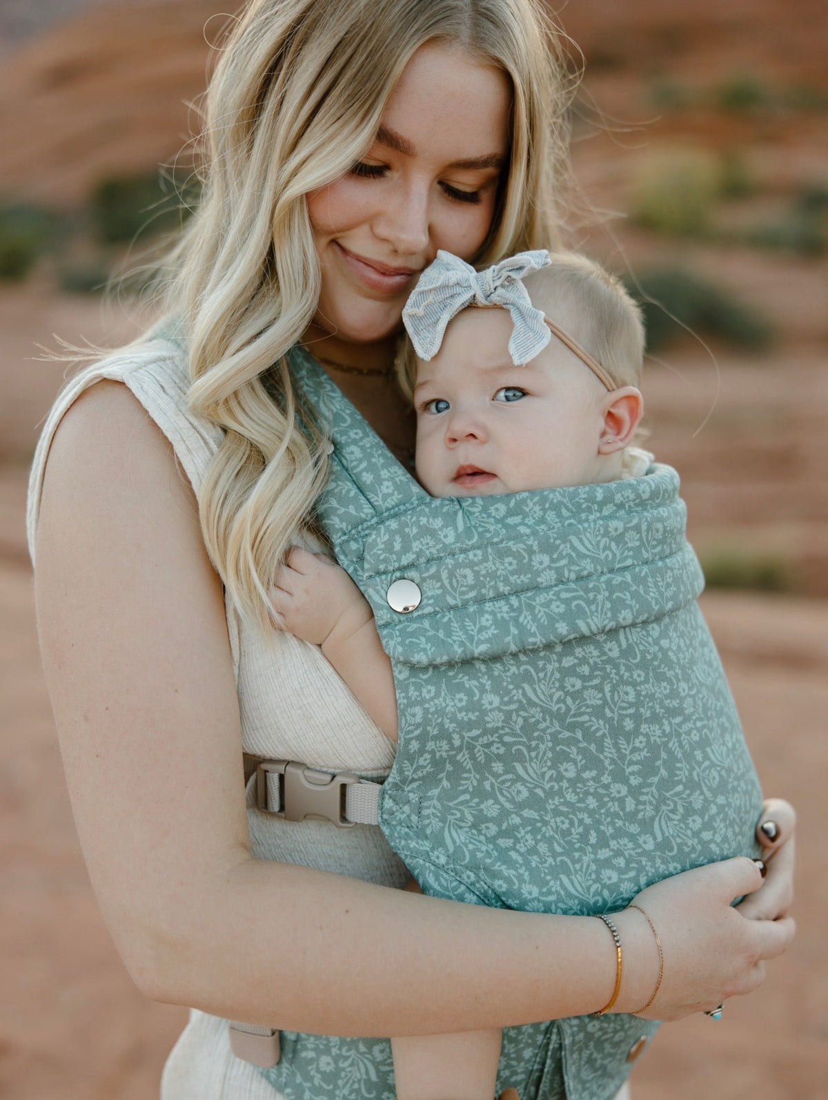 Teal floral baby carrier with pocket, worn by parent holding baby close