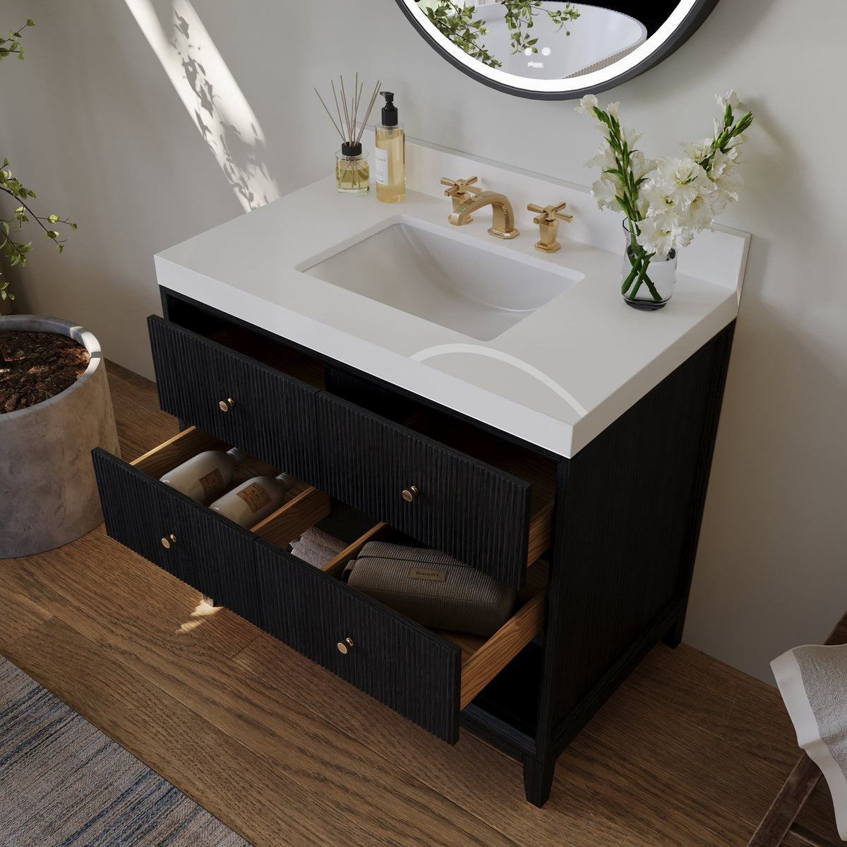 Black bath vanity with white sink and open drawers with gold knobs, round wall mirror above