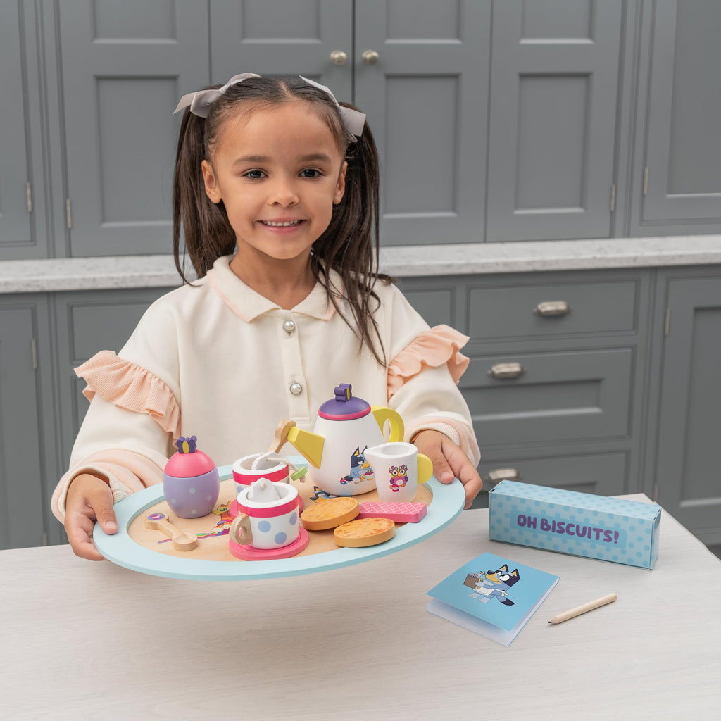 Child smiling with wooden tea set tray, teapot, cups, cookies, and card nearby