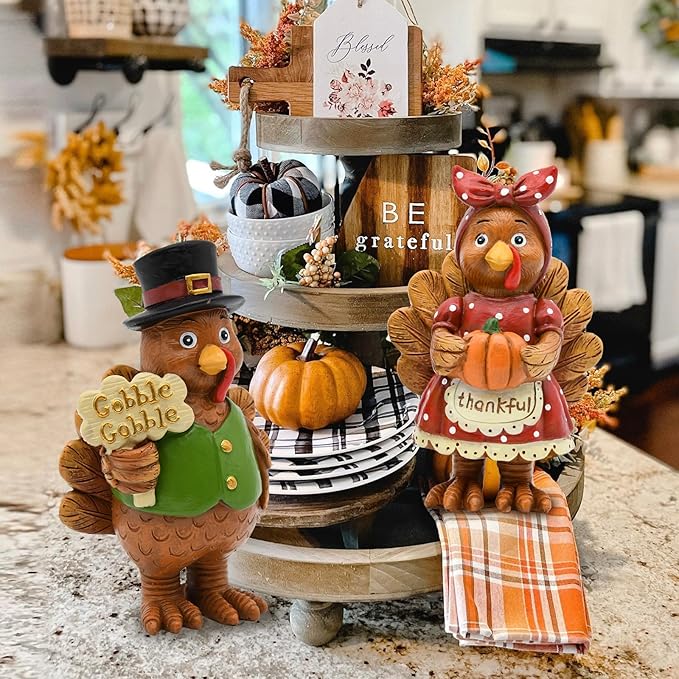 Resin turkey couple figurines standing on a table near pumpkins and fall decorations.
