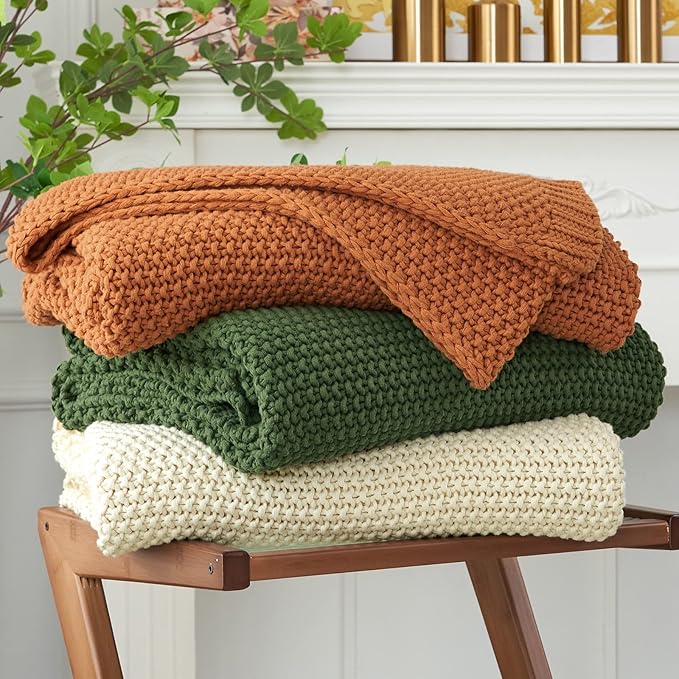 Chunky cable-knit throw blanket stack in olive green, cream, and rust-brown tones