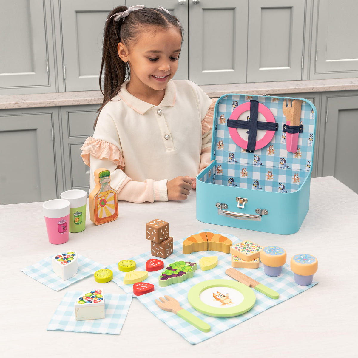 Wooden picnic basket set on table with child, drink cups, and play food items