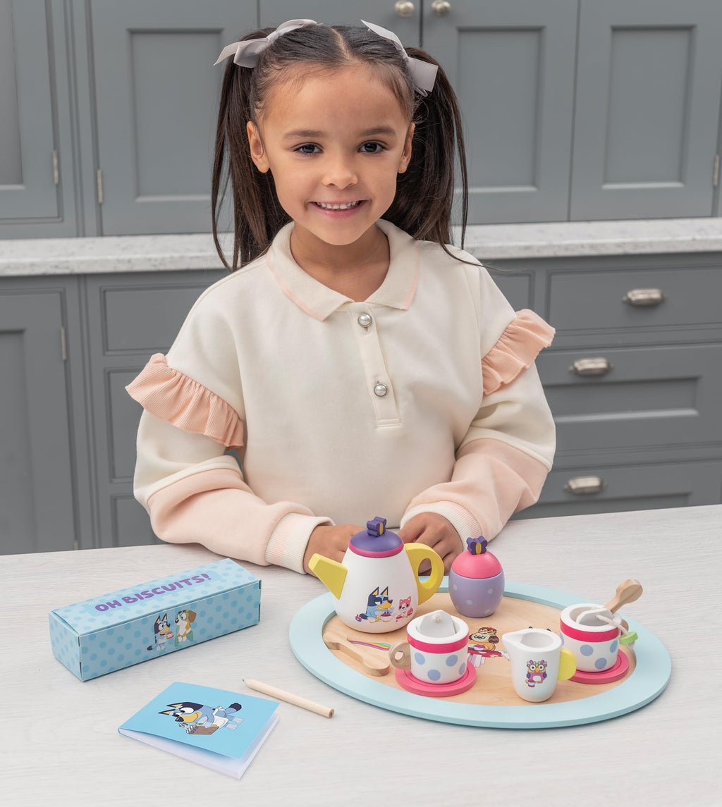 Child at table with wooden tea set: teapot, cups, saucers, cookies, and card