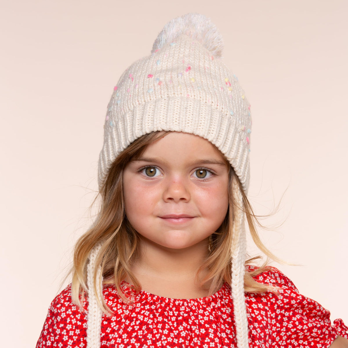 Off-white knit earflap beanie with pom-pom, tie strings, and small front brand patch.
